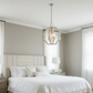 A modern pendant light with a silver-toned metal frame and clear glass panels hangs over a neatly made bed in a bright, neutral-toned bedroom.