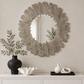 A round decorative wall mirror with a textured, scalloped wood-grain frame in a weathered gray finish, mounted on a beige wall above a white console table styled with vases and decor.
