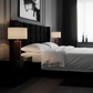 A modern bedroom scene featuring a black tufted bed frame with crisp white bedding, flanked by matching black nightstands and lit with warm table lamps. The background includes dark gray walls, a black headboard, and a window with sheer curtains.