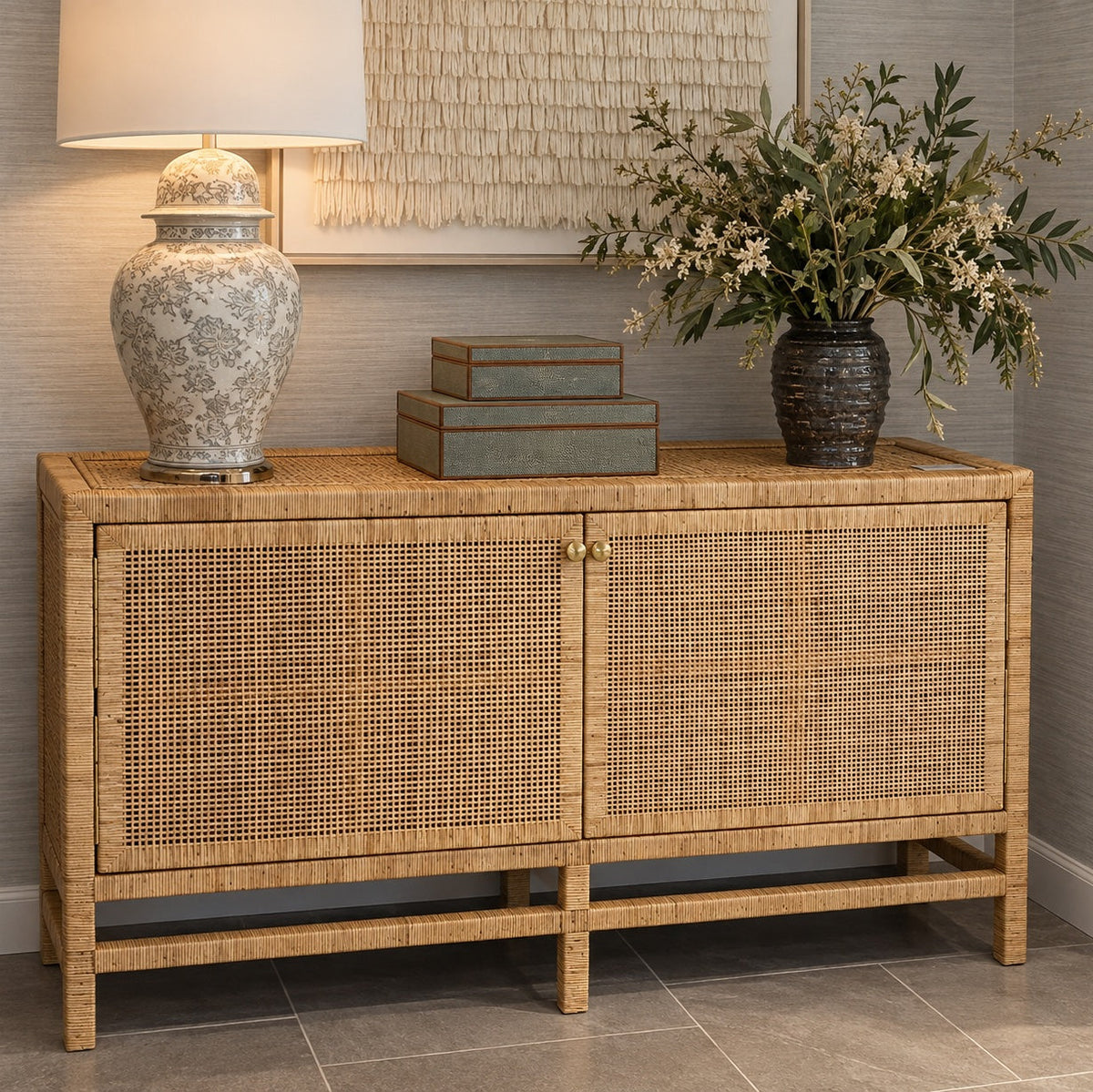 A mid-century modern sideboard cabinet made of natural woven rattan, styled with a ceramic table lamp, stacked boxes, and a floral arrangement on top.