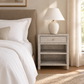 A light beige textured nightstand with a single drawer and woven shelf, styled in a bedroom next to a bed with white linens and a beige upholstered headboard. A matching table lamp sits on top.