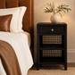 A black woven rattan nightstand with a single drawer and two open shelves, styled beside a bed with white linens and a brown leather headboard. A white ceramic vase with green branches sits on top.