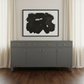 A modern gray sideboard cabinet with gold knobs, positioned against a white wall under a framed black abstract print, flanked by cream curtains and set on a dark wood floor.