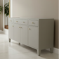 A modern sideboard cabinet in a light gray finish with textured drawer fronts and brass handles, set against a neutral beige wall and light marble floor.