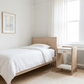 A minimalist bedroom scene featuring a light oak single bed with white bedding, a matching wooden nightstand with a white drawer, white curtains, and a white wall.