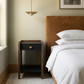 A modern bedside table with a dark wood frame, black drawer front, and brass pull, positioned next to a bed with white linens and a brown headboard in a softly lit bedroom.