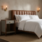 A styled bedroom scene featuring a bed with white bedding, a brown leather tufted headboard, a matching wooden nightstand, and wall-mounted sconces against a textured beige wall.