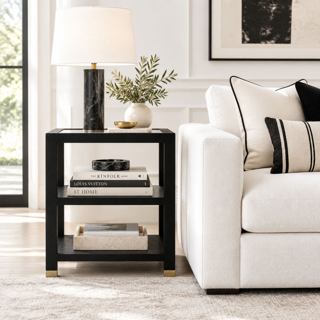 A modern black metal side table with gold-toned feet, styled in a bright living room next to a white sofa with decorative pillows.