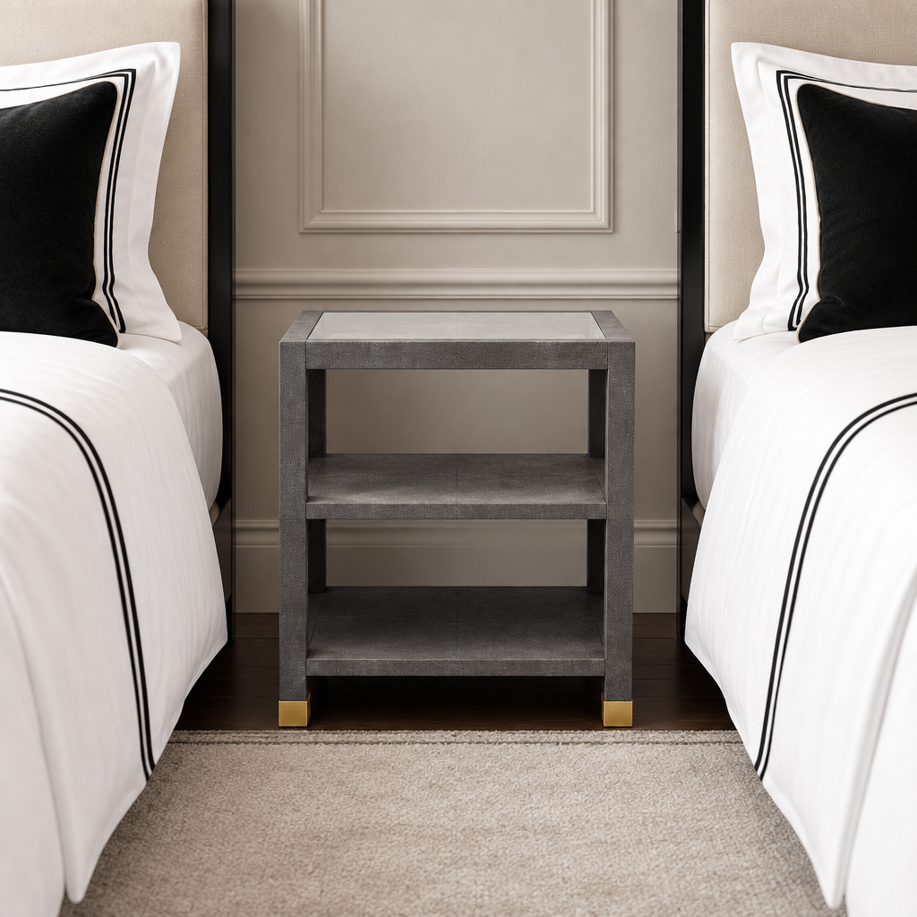A modern two-tier nightstand with a textured gray finish and gold metal feet, positioned between two matching beds with white linens and black pillows.