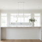 A modern kitchen island with a white subway tile base, a white marble countertop, and a white multi-light pendant chandelier hanging above. The kitchen features white walls, large multi-pane windows, and light wood flooring.