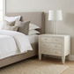 A light beige upholstered bed with white bedding stands beside a matching light wood nightstand with three drawers and a white table lamp, in a softly lit bedroom with a woven rug on the floor.