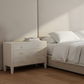 A light beige three-drawer nightstand in a minimalist bedroom setting, styled with a white vase of pampas grass and a stack of books next to a bed with white linens.