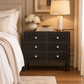 A black six-drawer nightstand with cream knobs and brass-toned trim, styled in a bedroom next to a bed with white linens. On top of the nightstand are a vase of white flowers, books, and a lit table lamp with a cream shade and marble base.