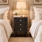 A dark charcoal three-drawer nightstand with silver knobs stands between two matching beds in a styled bedroom. A table lamp, a small vase of white flowers, and a candle rest on top of the nightstand.