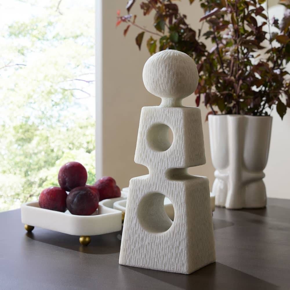 White abstract sculpture on a table with fruits and a plant in the background