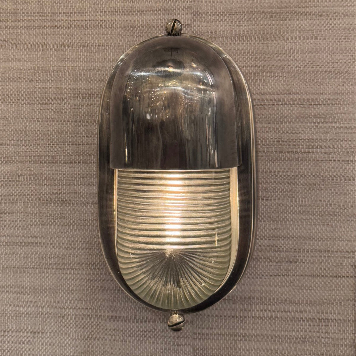 A vintage oval wall sconce with a polished silver metal frame and a ribbed clear glass shade, shown against a textured gray wall.