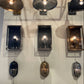 Three wall-mounted sconces displayed side by side on a white wall, each with a different finish: a dark bronze/black rectangular box, a silver rectangular box, and a bronze rectangular box, with matching dome-shaped shades above and oval lampshades below.