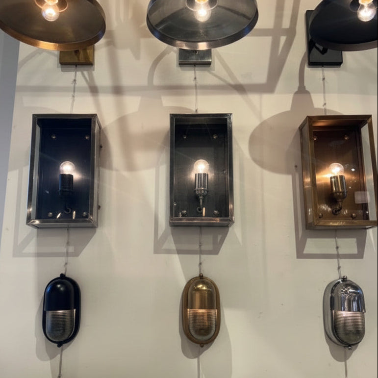 Three wall-mounted sconces displayed side by side on a white wall, each with a different finish: a dark bronze/black rectangular box, a silver rectangular box, and a bronze rectangular box, with matching dome-shaped shades above and oval lampshades below.