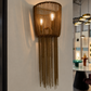A cylindrical pendant light fixture with a woven brown basket top and long dangling chain fringe, shown hanging on a white wall in a modern interior space.