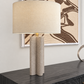 A table lamp with a large beige fabric drum shade and a textured, wavy rectangular concrete base, shown on a dark marble console table in front of a wall with framed art.
