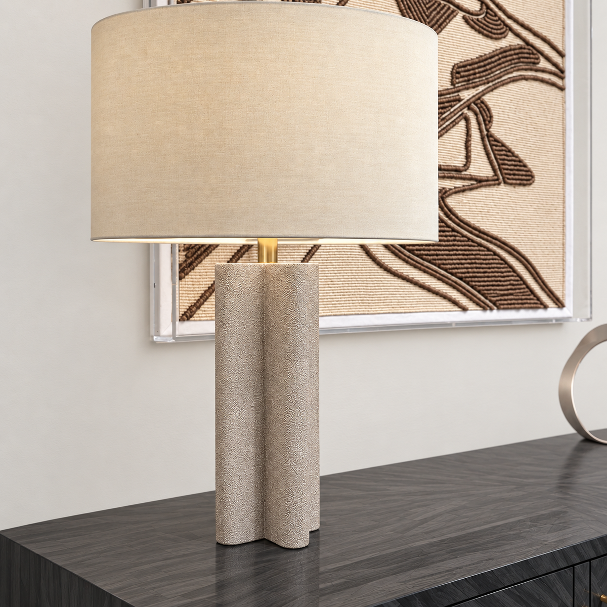 A table lamp with a large beige fabric drum shade and a textured, wavy rectangular concrete base, shown on a dark marble console table in front of a wall with framed art.