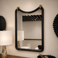 A rectangular wall mirror with a thick black frame and gold wall hooks at the top, mounted on a beige wall in a styled interior setting.