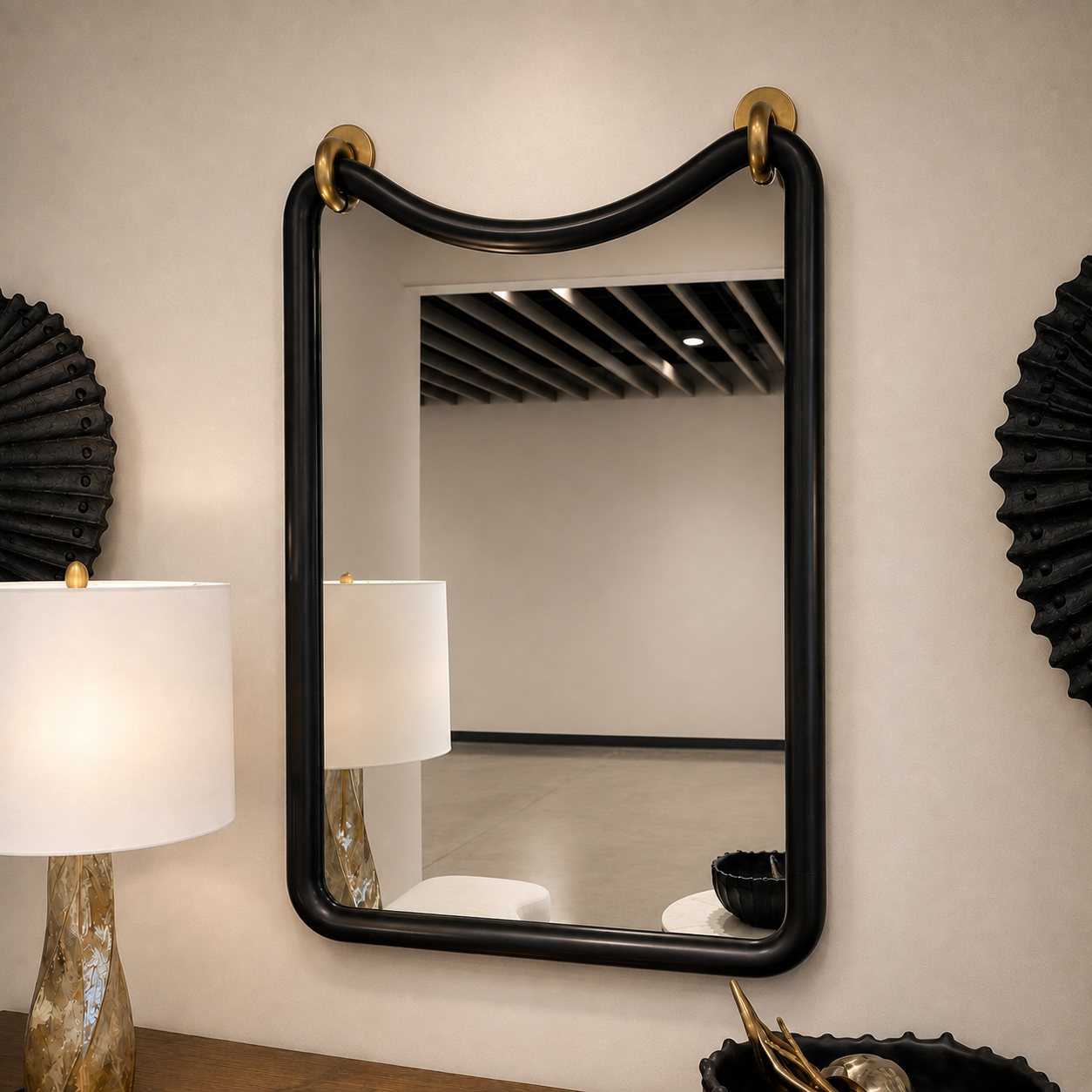 A rectangular wall mirror with a thick black frame and gold wall hooks at the top, mounted on a beige wall in a styled interior setting.