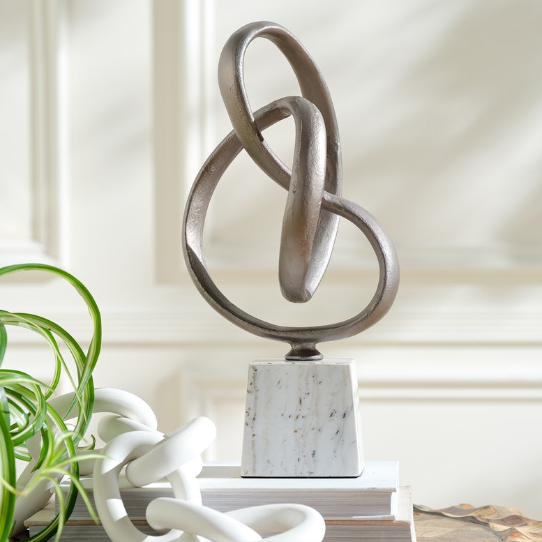 Decorative metal sculpture on a marble base with a plant and books in the background