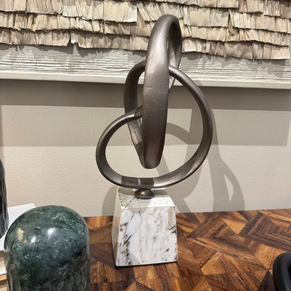 Abstract metal sculpture on a marble base against a shingled wall.