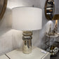 Table lamp with a white shade and silver base on a table against a tiled wall.