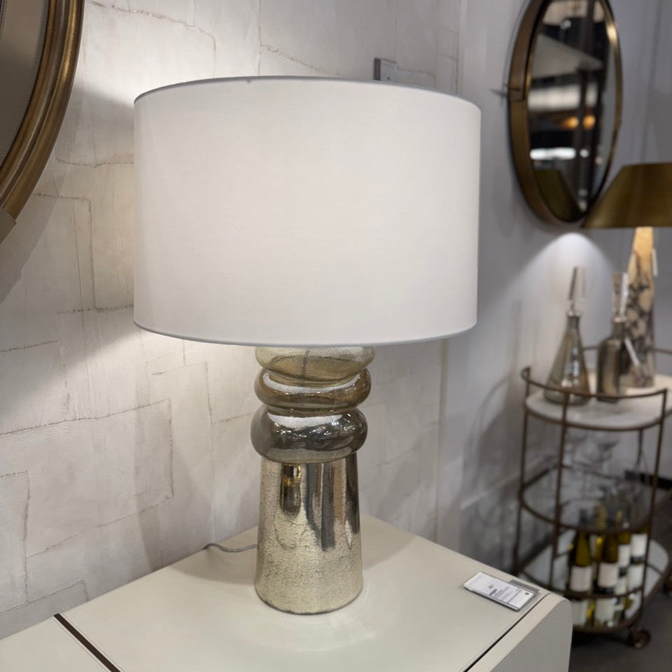 Table lamp with a white shade and silver base on a table against a tiled wall.