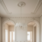 A bright, elegant living room with a large off-white sofa centered beneath a decorative chandelier, framed by two arched windows and detailed cream molding on the walls and ceiling.