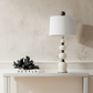 An off-white table lamp with a tapered drum shade and a stacked bead-like base with dark accents, styled on a white console table against a cream vertical slat wall.
