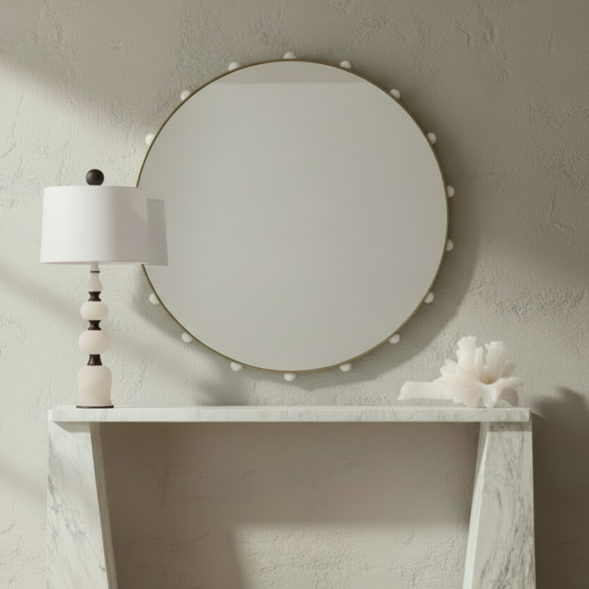 A round wall mirror with a beaded gold frame hangs above a white console table in a softly lit room. A white table lamp with a striped base stands on the right, and a white decorative object sits on the left.