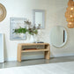 A natural woven rattan console table styled in a bright, modern entryway with a white vase of greenery, framed wall art, and multiple mirrors on a cream wall.
