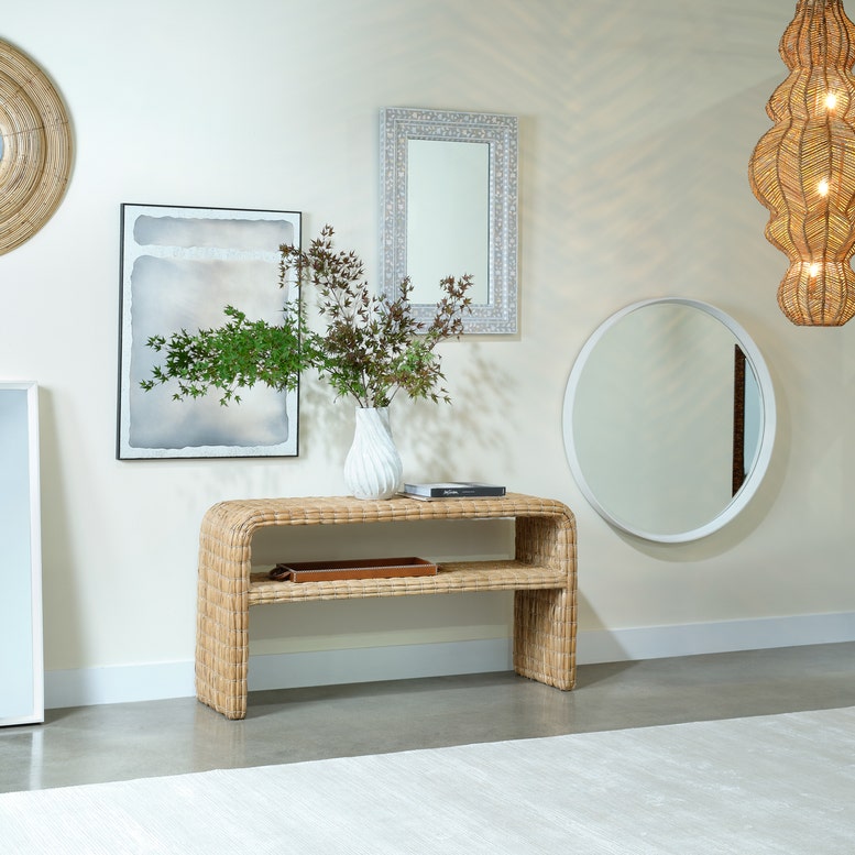 A natural woven rattan console table styled in a bright, modern entryway with a white vase of greenery, framed wall art, and multiple mirrors on a cream wall.