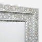 A close-up of a decorative rectangular mirror frame featuring a mother-of-pearl inlay of white and pale gold floral and leaf motifs on a light gray background.