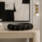A black ceramic ribbed bowl with gold trim sits on a light wood console table in a styled interior. A large black, white, and beige abstract painting hangs on the wall above, flanked by two gold wall sconces.