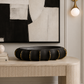A black ceramic bowl with gold trim sits on a light wood console table in a styled interior setting with a framed abstract print and wall sconce.