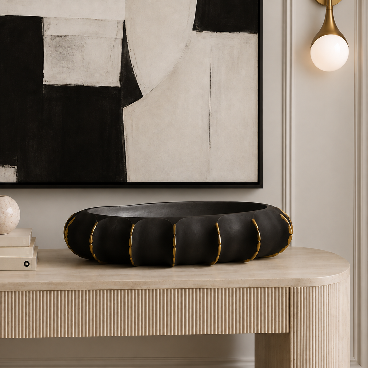 A black ceramic bowl with gold trim sits on a light wood console table in a styled interior setting with a framed abstract print and wall sconce.