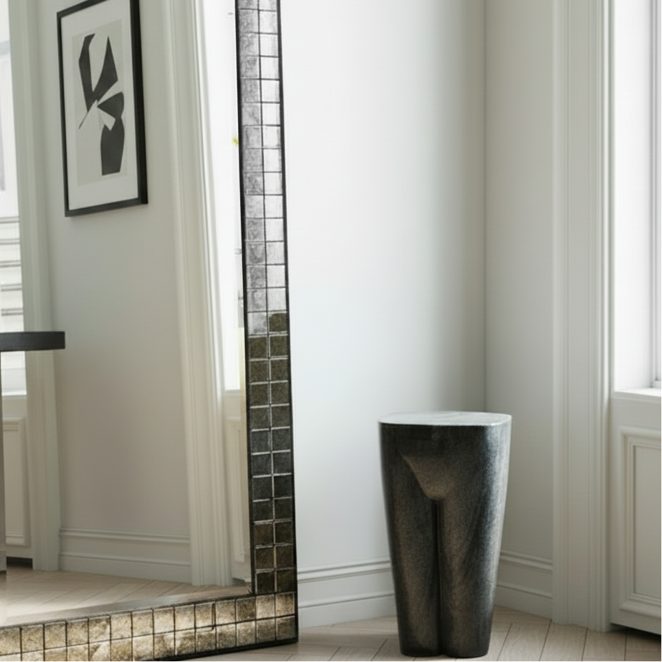 A tall rectangular floor mirror with a mosaic tile border, leaning against a white wall in a bright room. The mirror reflects a framed black-and-white artwork, and the scene includes a dark cylindrical stool and a window on the right.