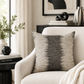 A square throw pillow with a textured black and beige ikat-style pattern, resting on a cream-colored armchair in a styled living room setting.