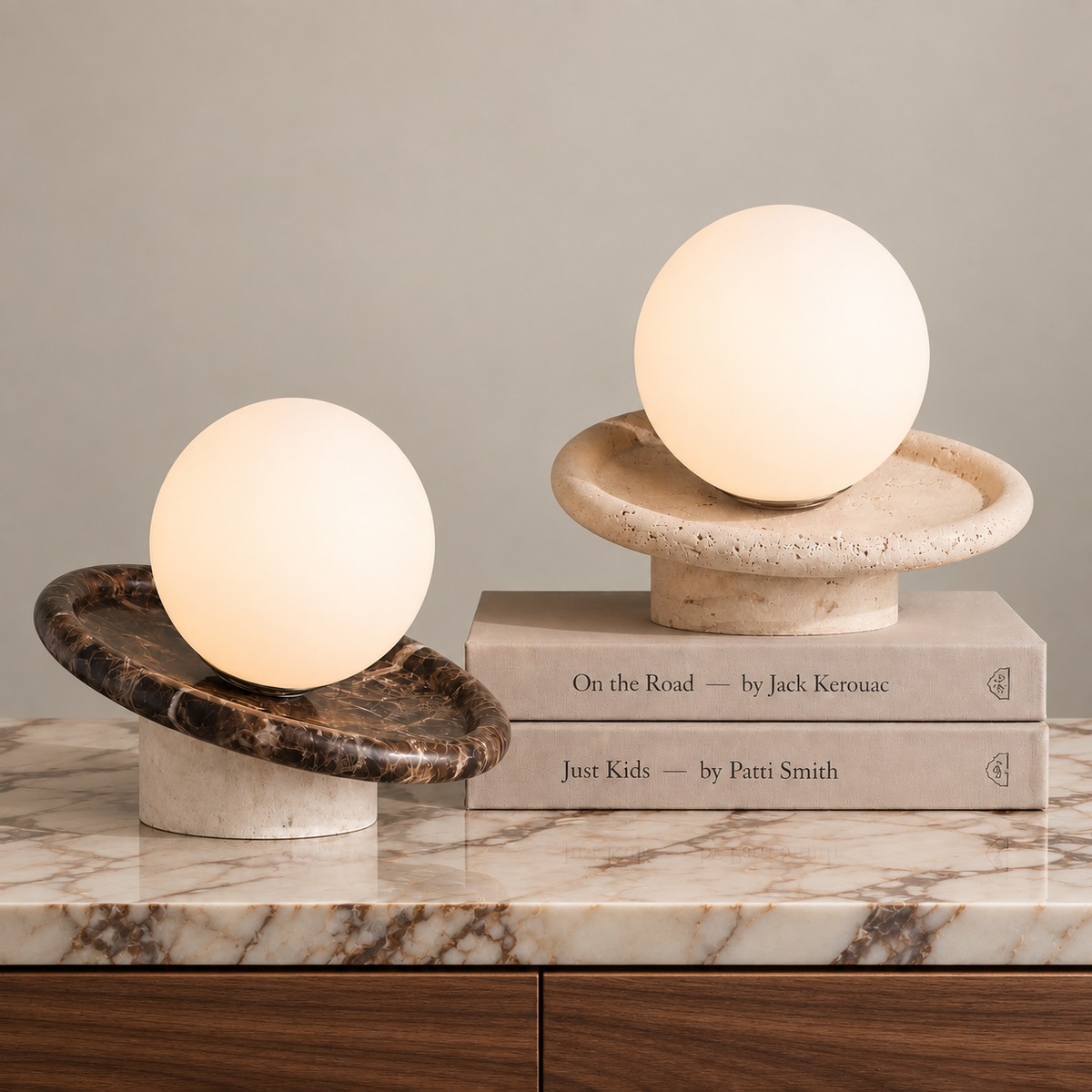 Two modern table lamps with glowing oval shades sit on a marble surface against a neutral wall. One lamp has a dark brown marble base, and the other is styled on stacked books with a light stone base.