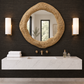 A round woven straw mirror with a gold-toned frame hangs on a dark textured wall above a white floating double-sink vanity with brass fixtures, flanked by two lit wall sconces.
