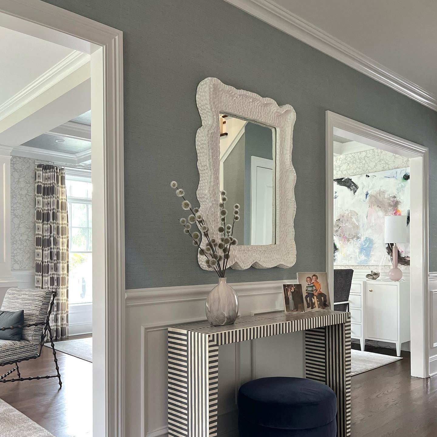Decorative console table with mirror and blue ottoman in a room with gray walls and wooden flooring.
