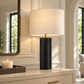 Norwood Table Lamp with a textured black sculptural base, brass accent, and ivory drum shade styled on a grasscloth console in a bright, elegant interior.