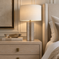Norwood Table Lamp styled on a cerused oak nightstand beside an upholstered bed, featuring a textured sculptural base, ivory drum shade, and warm brass accents.