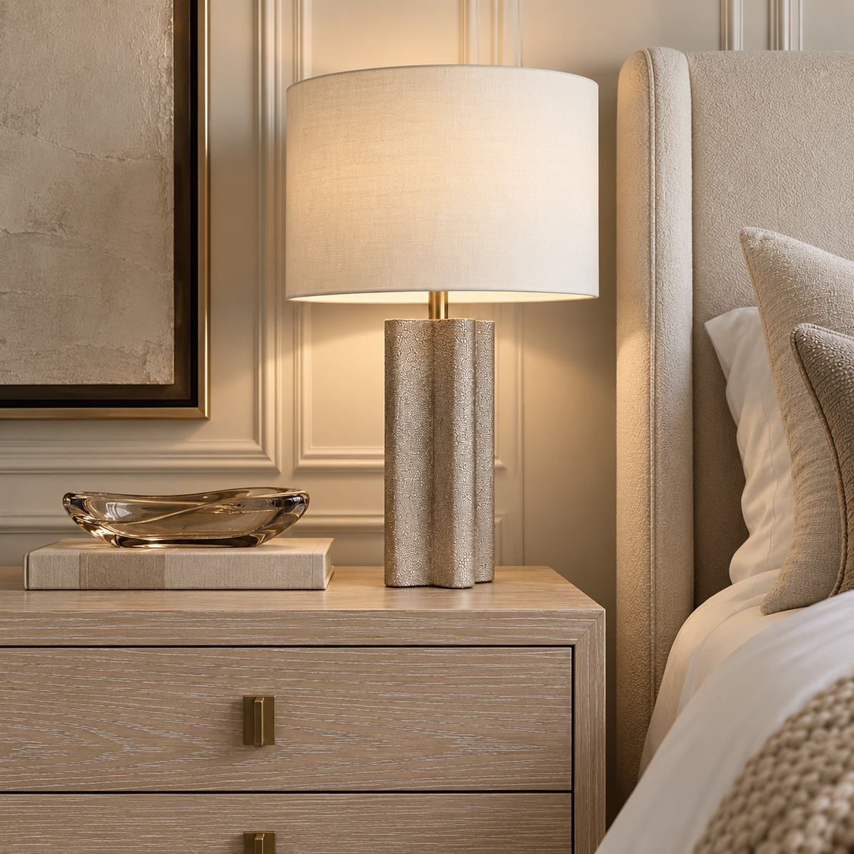 Norwood Table Lamp styled on a cerused oak nightstand beside an upholstered bed, featuring a textured sculptural base, ivory drum shade, and warm brass accents.