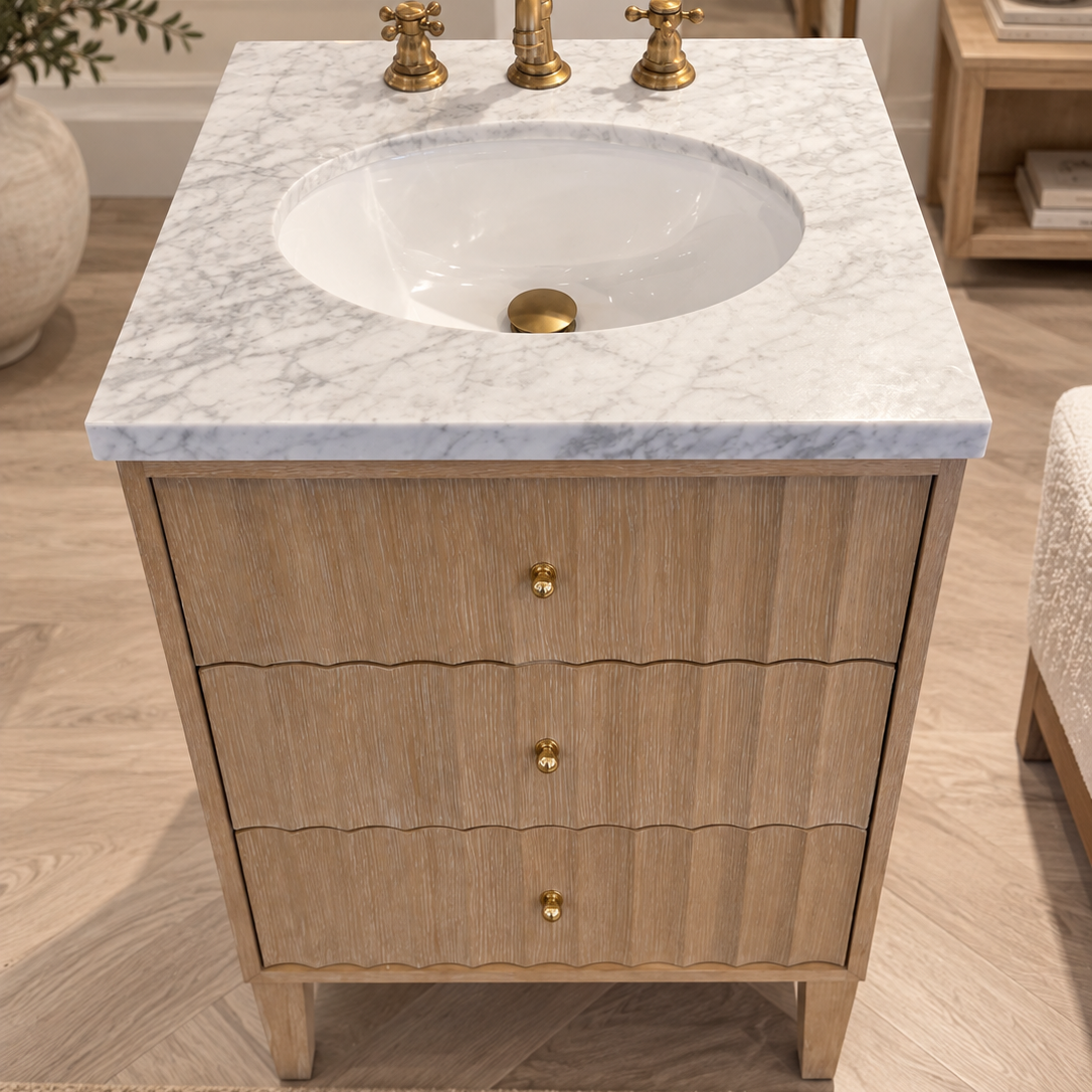 A single-sink bathroom vanity with a light natural wood cabinet, three drawers with scalloped fronts, and a white marble top with subtle gray veining. The vanity is equipped with a white oval basin and a polished gold-tone faucet set, including a central pedestal spout and two separate handles.