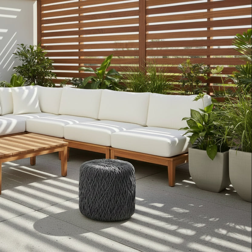 A dark gray textured pouf ottoman with a thick braided rope-like fabric cover, shown on a sunny patio